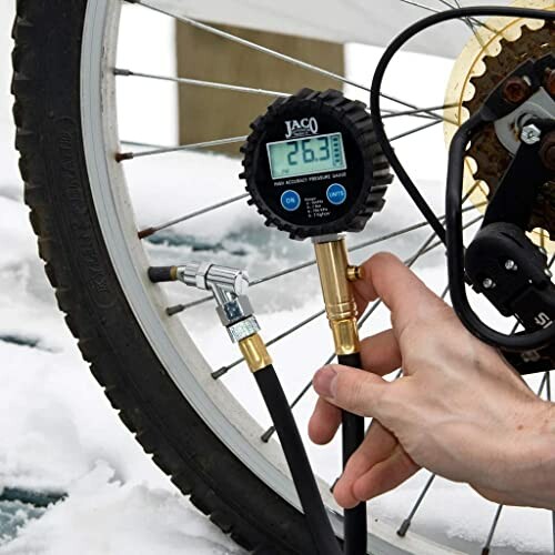 Person checking bicycle tire pressure with digital gauge in snowy setting.