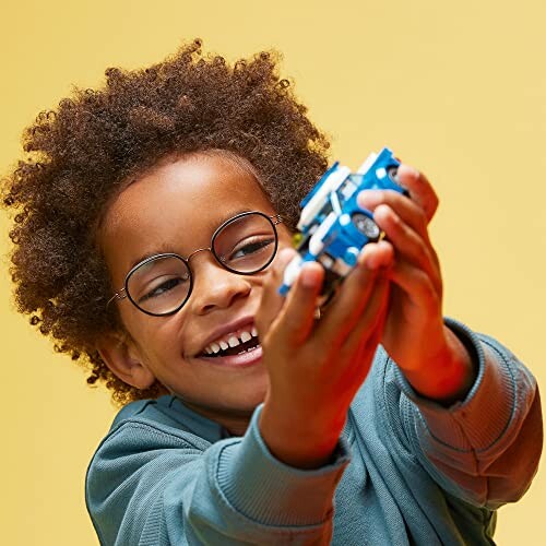 Child with glasses smiling and holding a blue toy car against a yellow background.