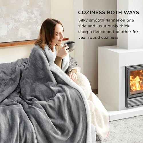 Woman wrapped in a gray blanket sitting by a fireplace with a mug.